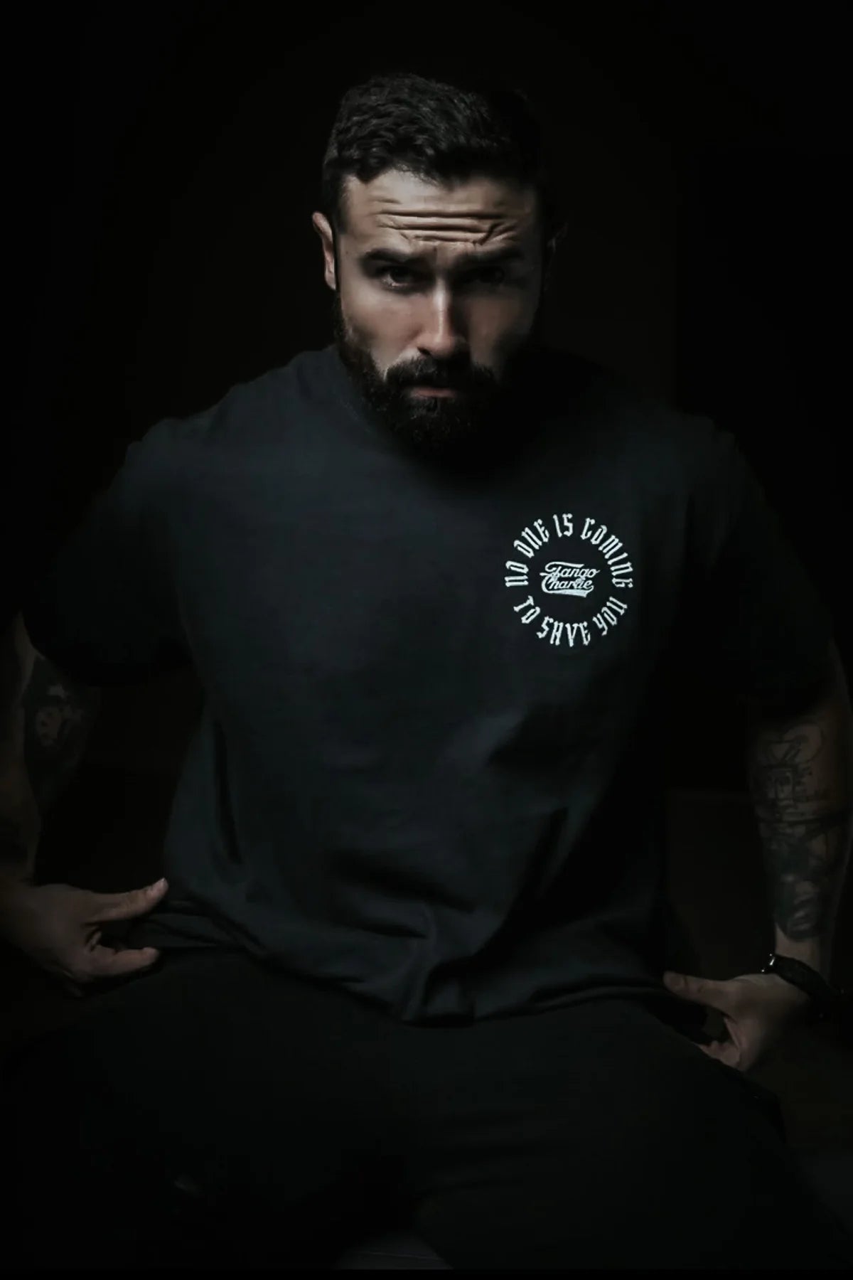 A bearded, tattooed man sits in a dark setting wearing the "No One Is Coming To Save You Tee - Black" by Tango Charlie Apparel. Light emphasizes his serious expression and the shirt’s circular white logo, while the background is shadowed.