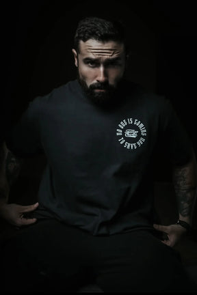 A bearded, tattooed man sits in a dark setting wearing the "No One Is Coming To Save You Tee - Black" by Tango Charlie Apparel. Light emphasizes his serious expression and the shirt’s circular white logo, while the background is shadowed.