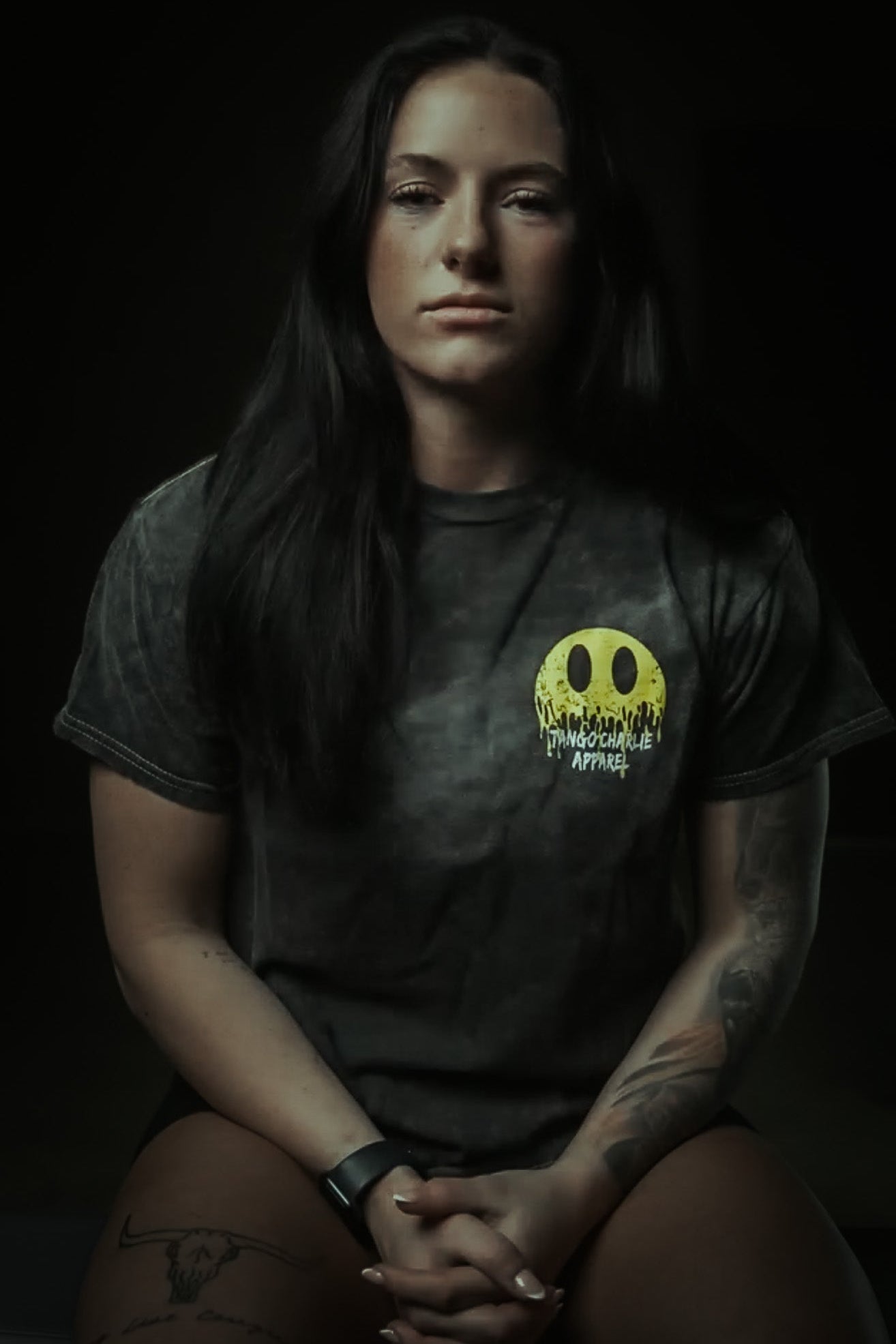A woman with long dark hair and arm tattoos sits with her hands clasped, wearing a Tango Charlie Apparel "Unleash The Savage Premium Tee" featuring a yellow melting smiley face logo. She also wears a wristwatch.