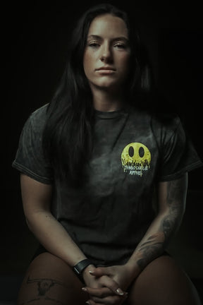 A woman with long dark hair and arm tattoos sits with her hands clasped, wearing a Tango Charlie Apparel "Unleash The Savage Premium Tee" featuring a yellow melting smiley face logo. She also wears a wristwatch.