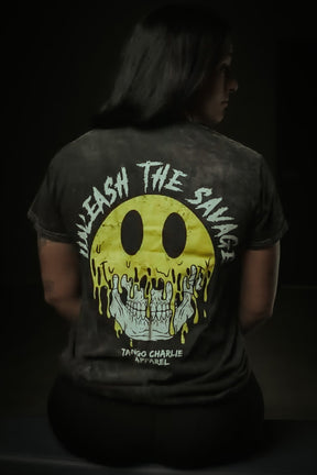 Someone sits facing away, wearing the Unleash The Savage Premium Tee by Tango Charlie Apparel, featuring a melting yellow smiley revealing skulls and edgy text. A dim background highlights the shirt's bold vibe.