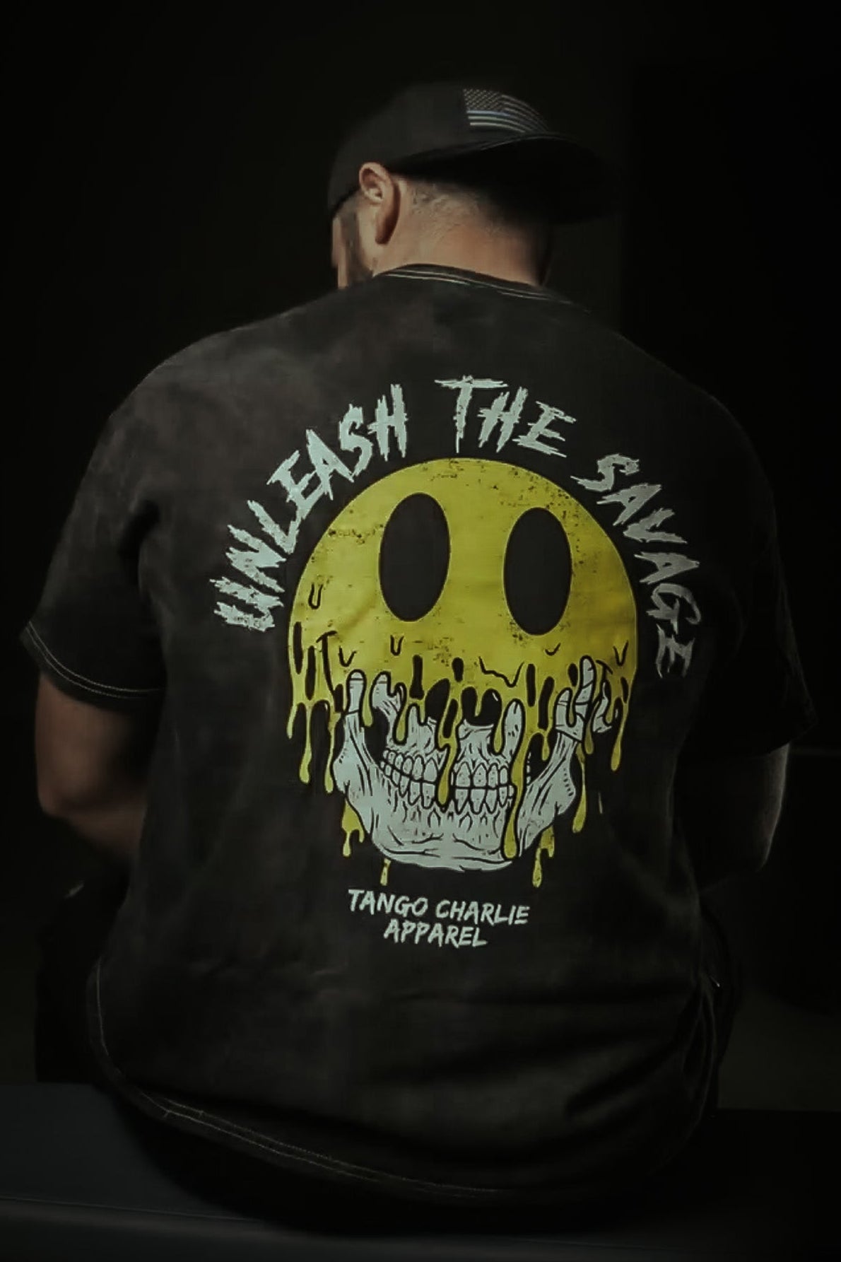 Wearing a dark cap and the Unleash The Savage Premium Tee by Tango Charlie Apparel, featuring a bold yellow melting smiley face and skull graphic. The back displays "UNLEASH THE SAVAGE" and "TANGO CHARLIE APPAREL.