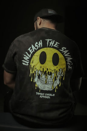 Wearing a dark cap and the Unleash The Savage Premium Tee by Tango Charlie Apparel, featuring a bold yellow melting smiley face and skull graphic. The back displays "UNLEASH THE SAVAGE" and "TANGO CHARLIE APPAREL.