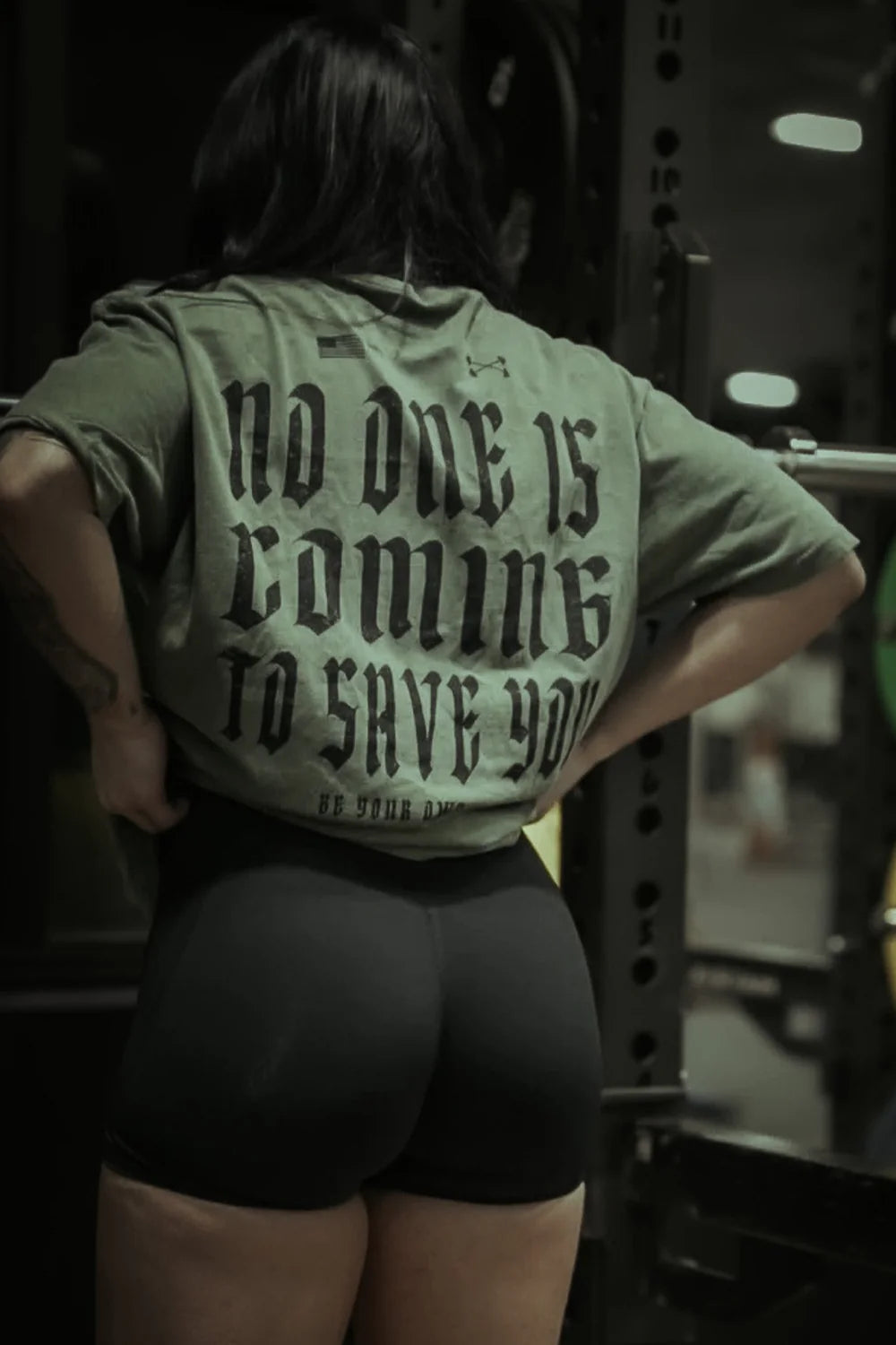A person stands in a gym wearing the Tango Charlie Apparel No One Is Coming To Save You Tee–Hemp, ready to take charge of their workout. The green shirt’s bold message inspires action as they prepare by weightlifting equipment.