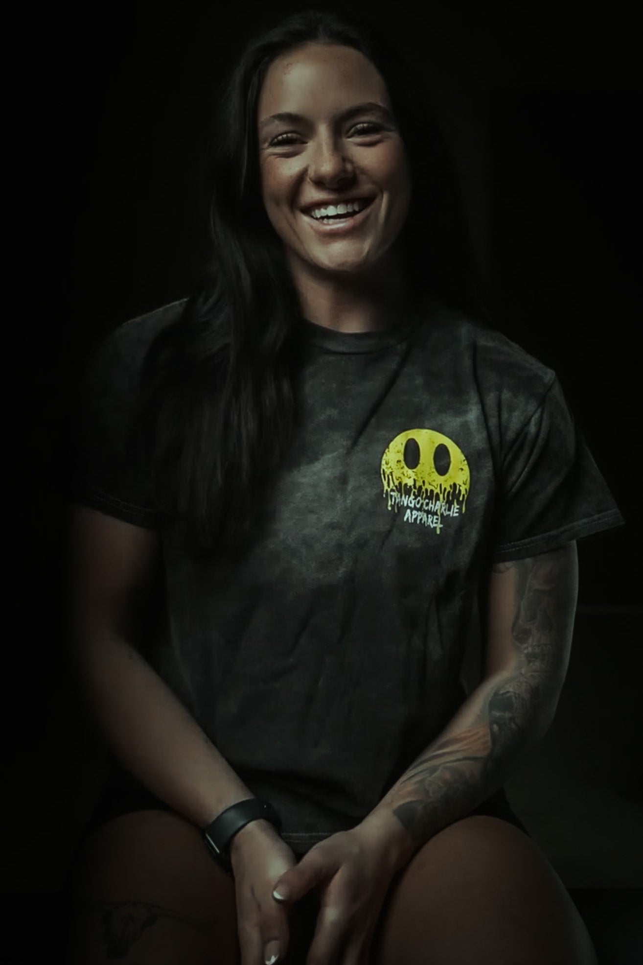 A woman with long dark hair smiles as she sits, wearing the Unleash The Savage Premium Tee by Tango Charlie Apparel, featuring a yellow melting smiley face and tattoos on her left arm, set against a dark background.