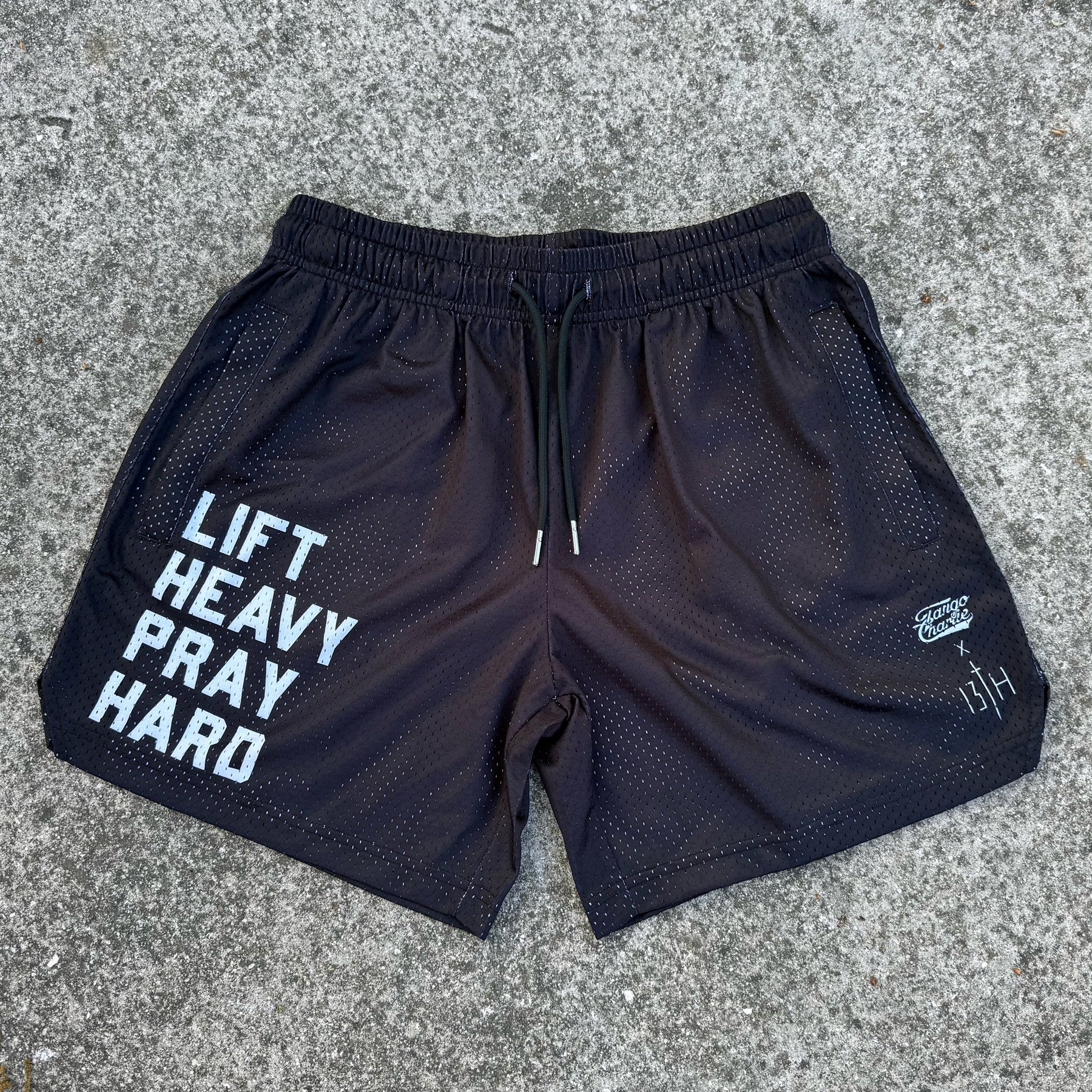 Tango Charlie Apparel's Men's Mesh Lifestyle Shorts | 5.5" - Lift Heavy Pray Hard Black feature bold white "LIFT HEAVY PRAY HARD" text on the left leg, small white logos on the right, and are shown on a rugged concrete surface—ideal for active living.