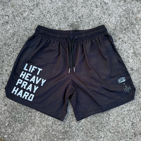 Tango Charlie Apparel's Men's Mesh Lifestyle Shorts | 5.5" - Lift Heavy Pray Hard Black feature bold white "LIFT HEAVY PRAY HARD" text on the left leg, small white logos on the right, and are shown on a rugged concrete surface—ideal for active living.