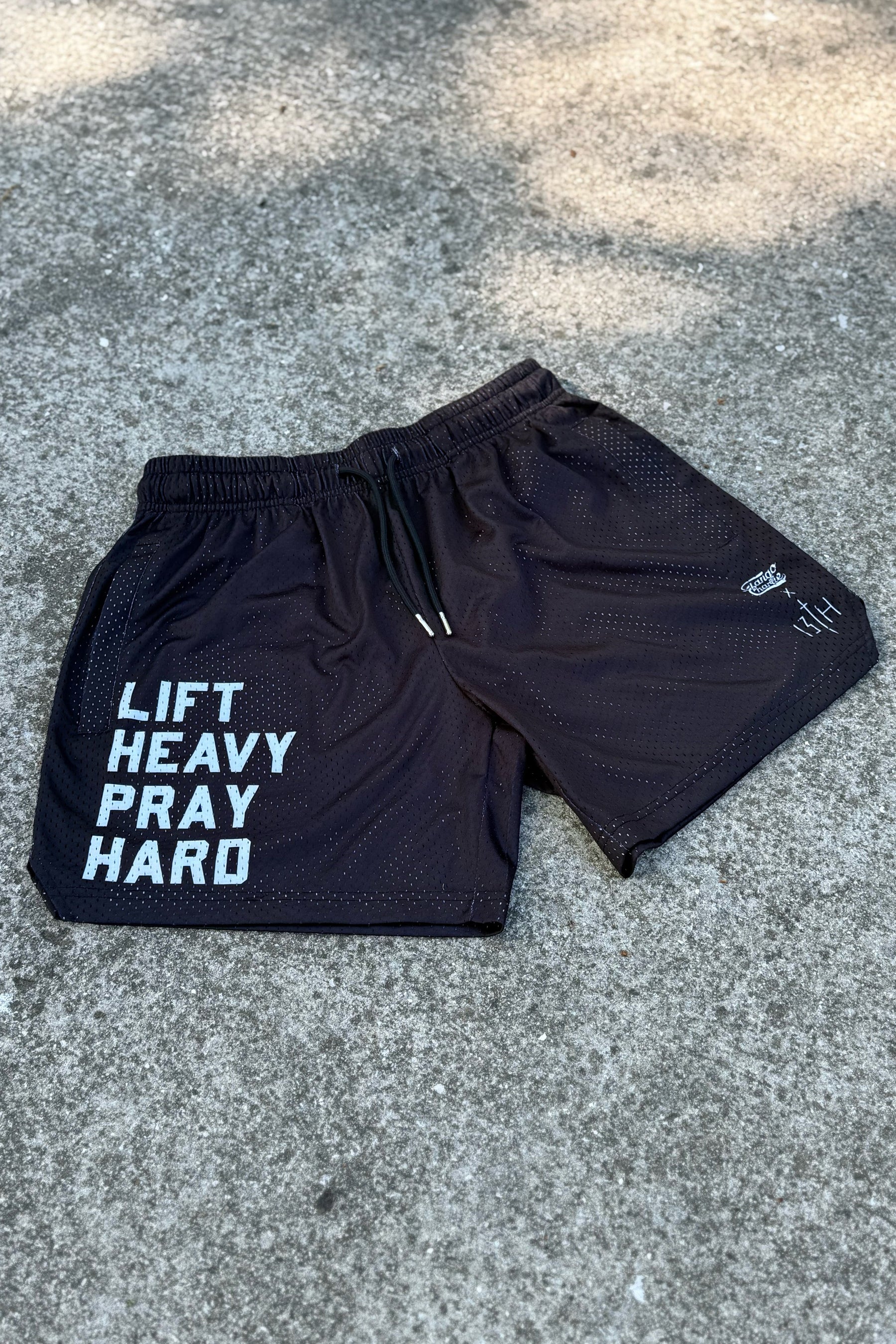 The Men's Mesh Lifestyle Shorts | 5.5"- Lift Heavy Pray Hard Black by Tango Charlie Apparel are black gym shorts featuring "LIFT HEAVY PRAY HARD" in white on the left leg, shown laid flat on concrete.