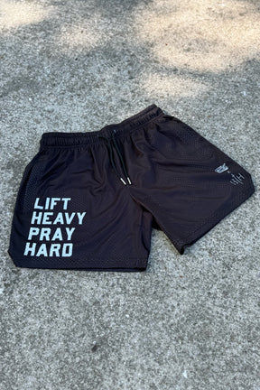 The Men's Mesh Lifestyle Shorts | 5.5"- Lift Heavy Pray Hard Black by Tango Charlie Apparel are black gym shorts featuring "LIFT HEAVY PRAY HARD" in white on the left leg, shown laid flat on concrete.