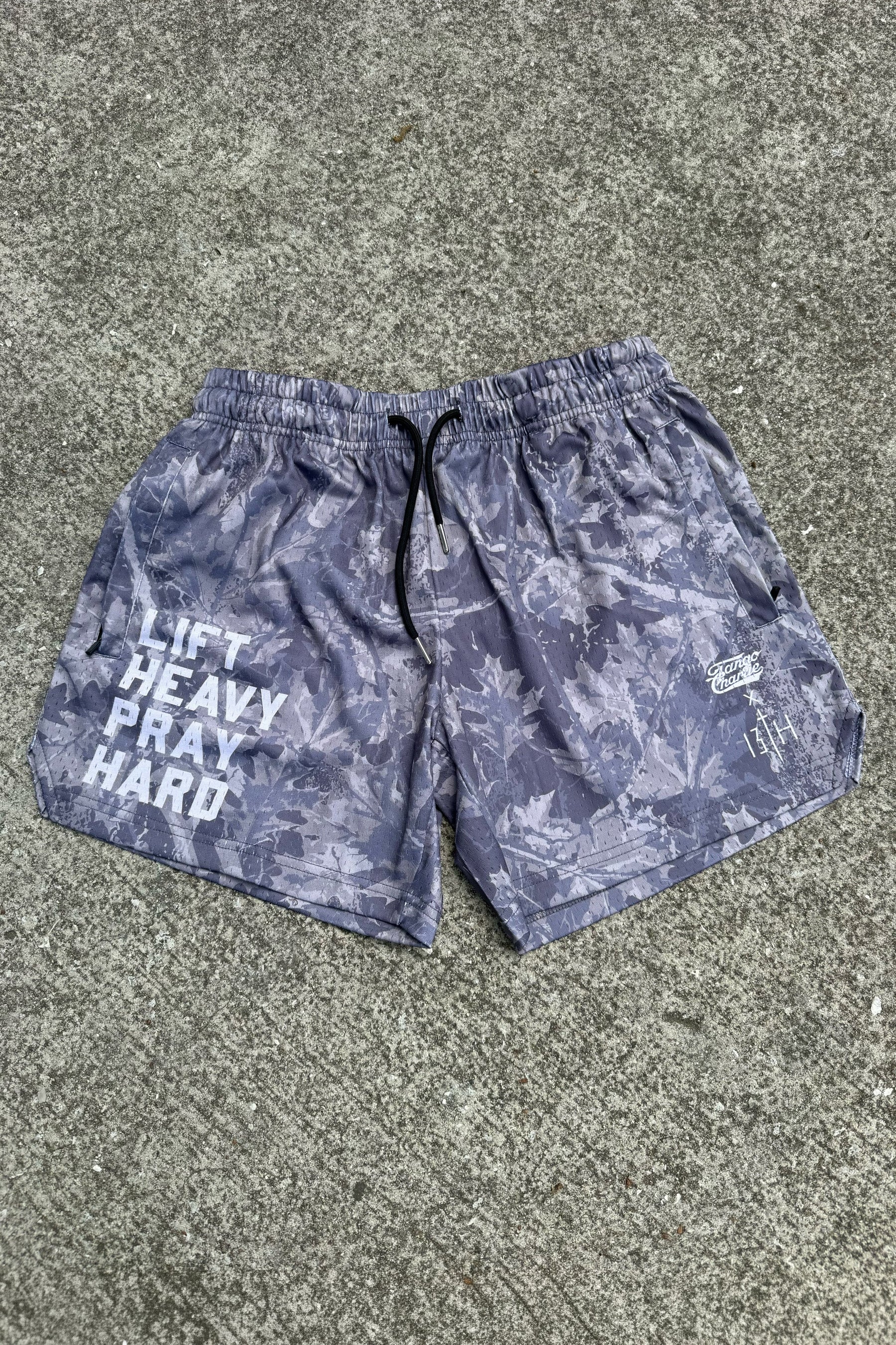 Tango Charlie Apparel Men's Mesh Lifestyle Shorts | 5.5" - Lift Heavy Pray Hard Deadwood, gray with leaf pattern and “LIFT HEAVY PRAY HARD” in bold white on the left leg, displayed on a concrete surface.