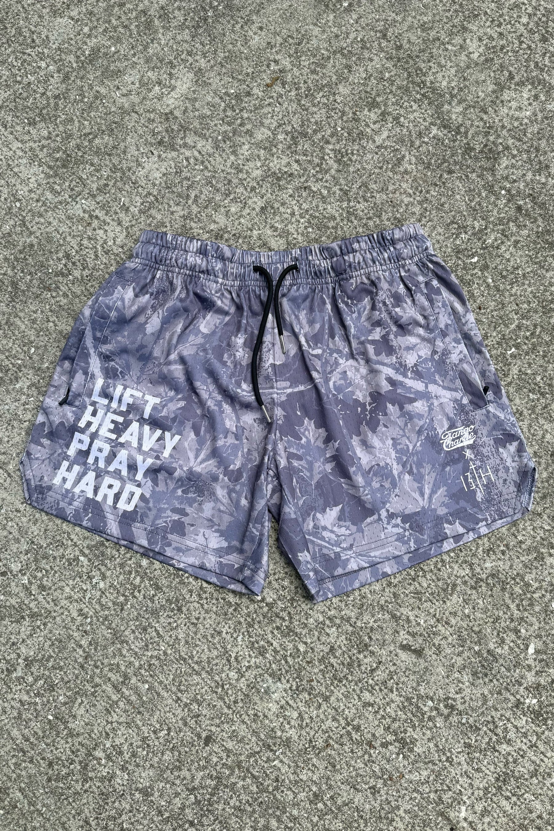 The Tango Charlie Apparel Men's Mesh Lifestyle Shorts (5.5") in gray feature a leaf pattern, "LIFT HEAVY PRAY HARD" text on the left leg, and a small logo on the right leg, shown lying flat on concrete.