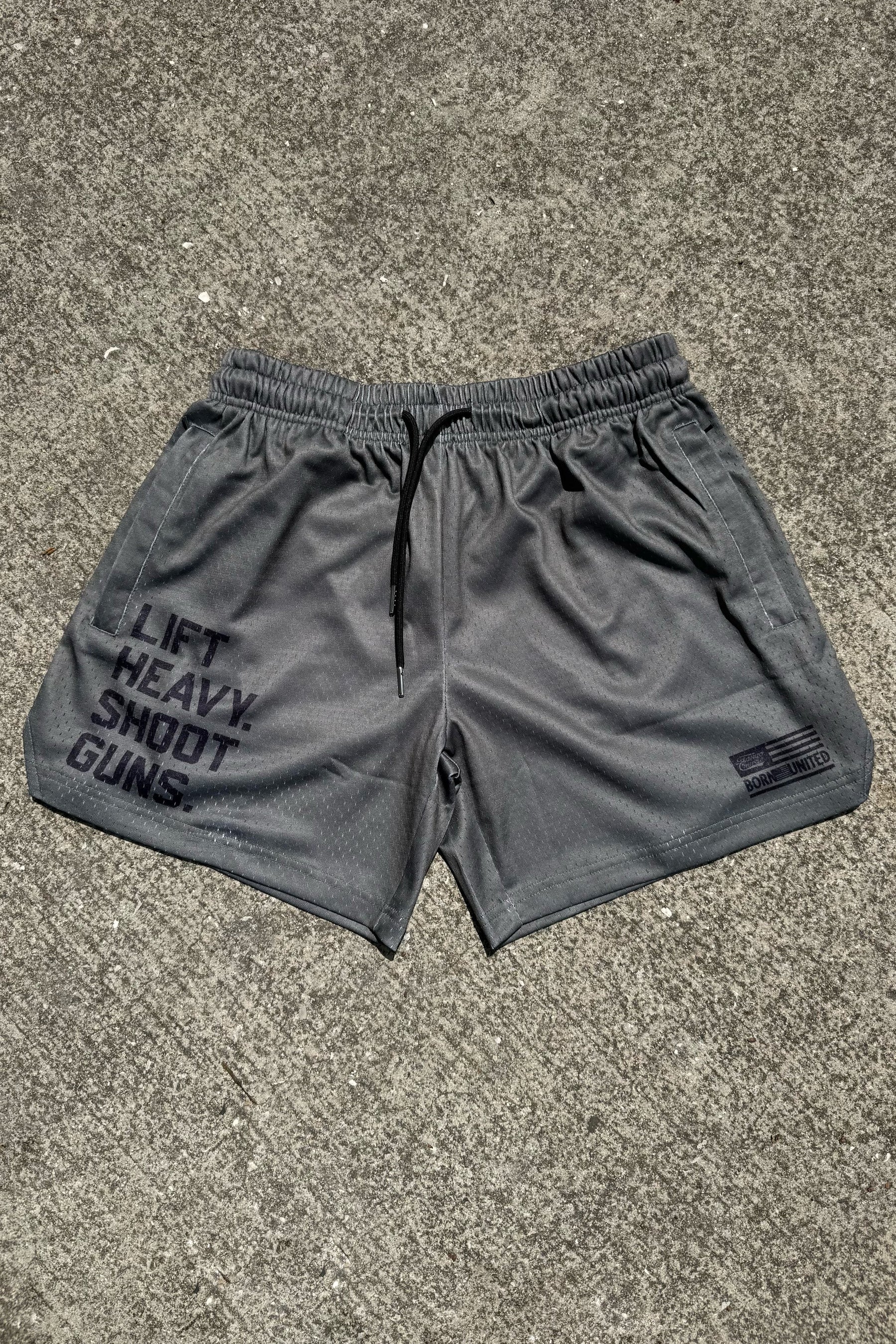 Tango Charlie Apparel Men's Mesh Lifestyle Shorts in OD Green with black drawstrings and a 5.5" inseam rest on concrete. The left leg reads LIFT HEAVY. SHOOT GUNS., while the right features a small logo and added text—ideal for active days.