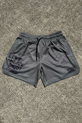 Tango Charlie Apparel Men's Mesh Lifestyle Shorts in OD Green with black drawstrings and a 5.5" inseam rest on concrete. The left leg reads LIFT HEAVY. SHOOT GUNS., while the right features a small logo and added text—ideal for active days.