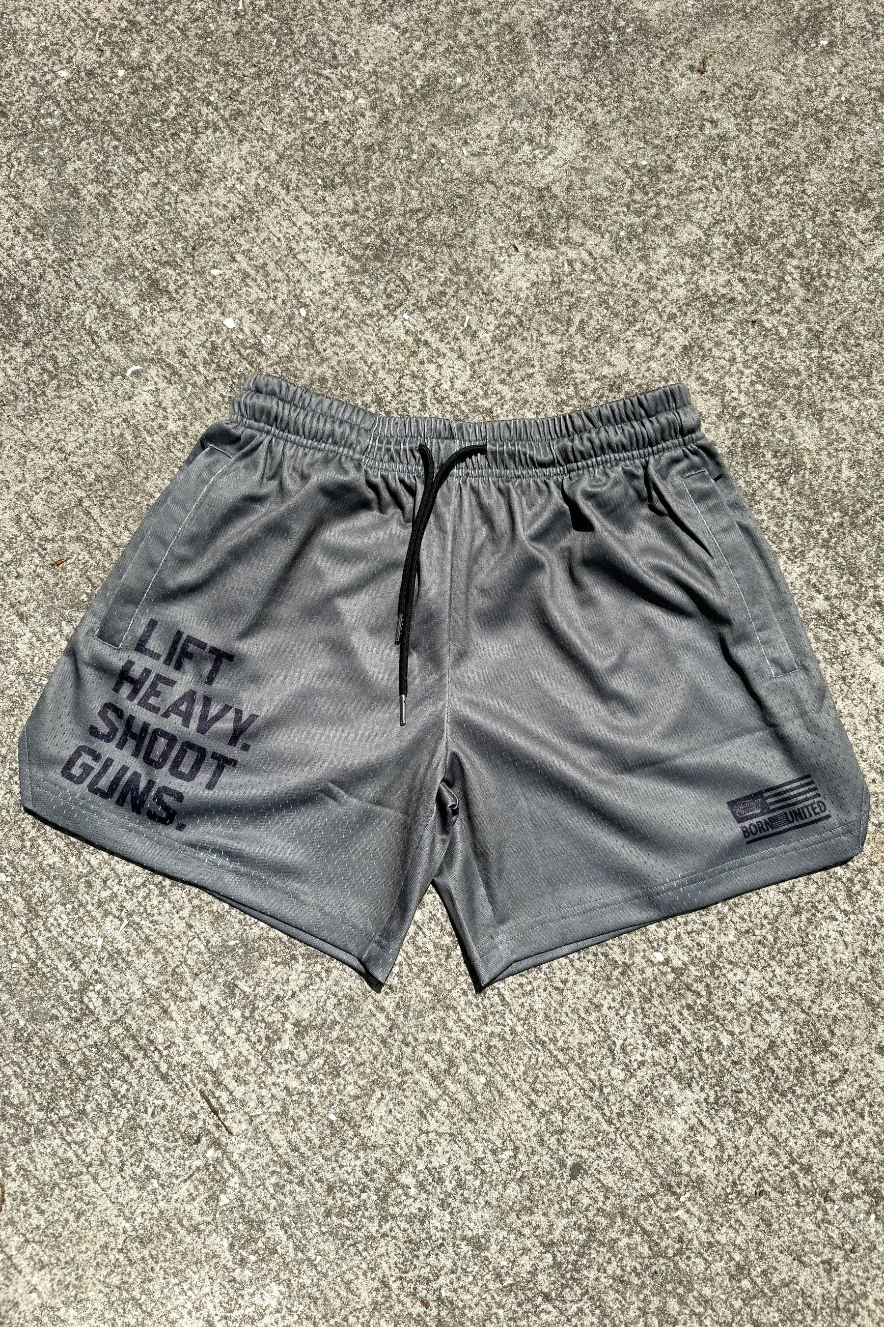 Tango Charlie Apparel's Men's Mesh Lifestyle Shorts in OD Green with a 5.5" inseam feature "LIFT HEAVY, SHOOT GUNS" on the left leg and a small NOBLE UNITED logo on the right, displayed on a concrete surface.