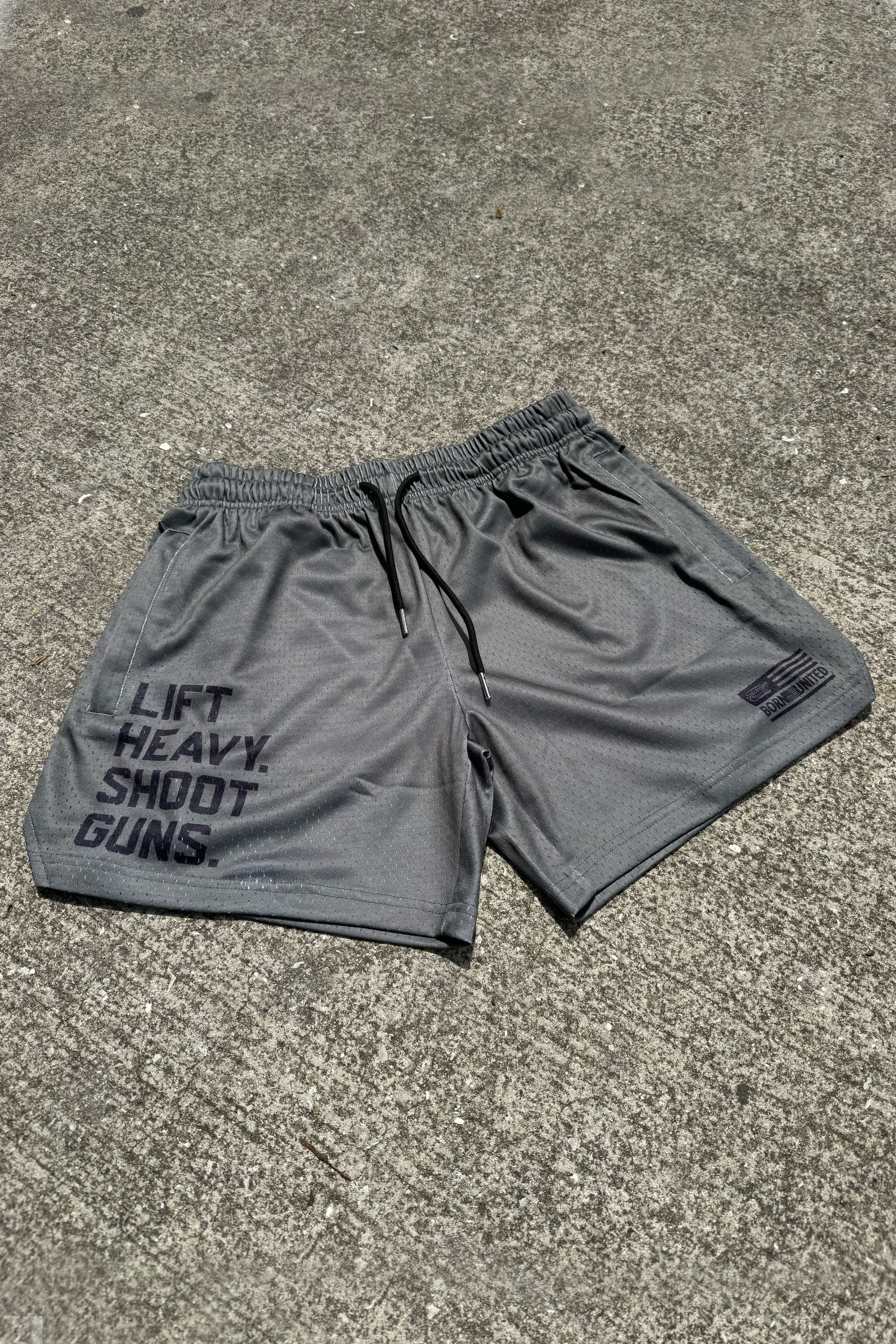 A pair of Tango Charlie Apparel Men's Mesh Lifestyle Shorts | 5.5"- LHSG in OD Green, featuring a drawstring waistband, rests on concrete with “LIFT HEAVY. SHOOT GUNS.” printed in bold black on the left leg.