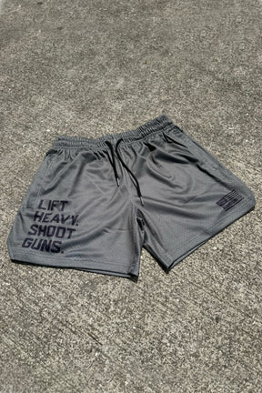 A pair of Tango Charlie Apparel Men's Mesh Lifestyle Shorts | 5.5"- LHSG in OD Green, featuring a drawstring waistband, rests on concrete with “LIFT HEAVY. SHOOT GUNS.” printed in bold black on the left leg.