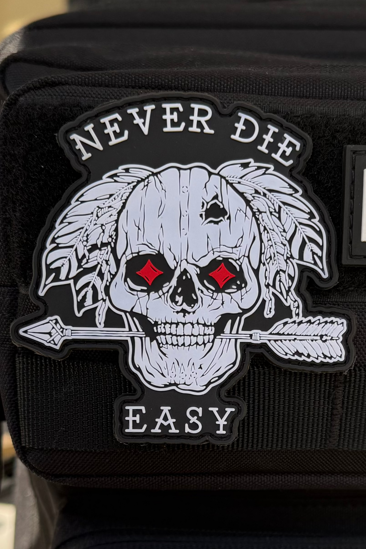 The Never Die Easy PVC Patch by Relentless Betrayal features a striking skull with red eyes, feathers, and an arrow through it, with the words NEVER DIE EASY above and below—a legacy-inspired design perfect for adding bold character to your gear.