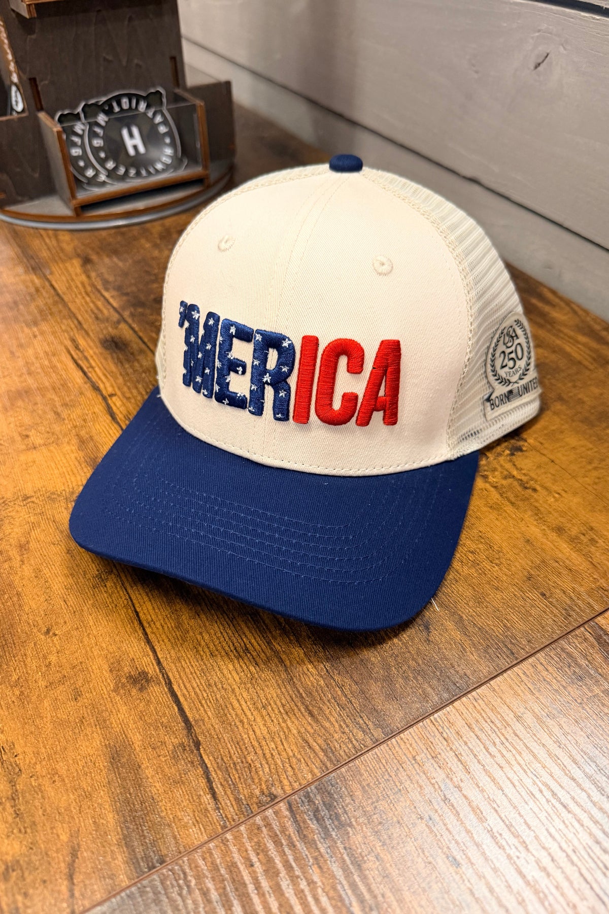 The Born United Private Label ’Merica Trucker Snapback is a white and blue trucker hat featuring “MERICA” embroidered in stars, stripes, and red across the front. Resting on a wooden surface, this handmade cap includes a breathable snapback enclosure for all-day comfort and a side patch reading “250 Commemorative Edition.”.