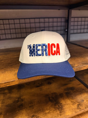 The Born United Private Label ’Merica Trucker Snapback showcases a white front, blue brim, and “MERICA” embroidered in bold red letters with star-and-stripe detailing. Displayed on a wooden shelf, this handmade hat features a breathable snapback enclosure for all-day comfort and timeless style.