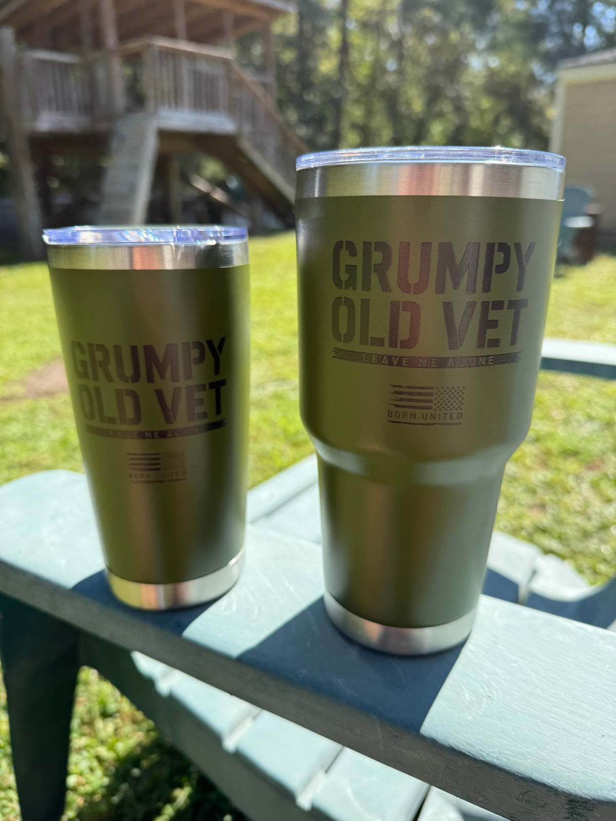 Two green "Grumpy Old Vet Tumbler - 20oz" by Born United Private Label rest on an outdoor table, with sunlight highlighting a wooden structure and grassy yard in the background.
