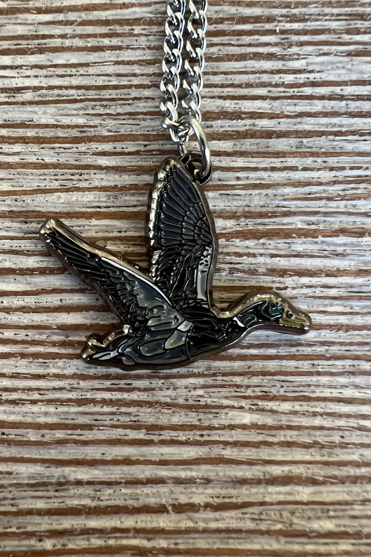 The Duck It OG Necklace by Mucker Bucket features a stainless steel flying bird pendant with detailed wings and body, set against a textured wood background and finished with a shiny surface. Proudly made in the USA.
