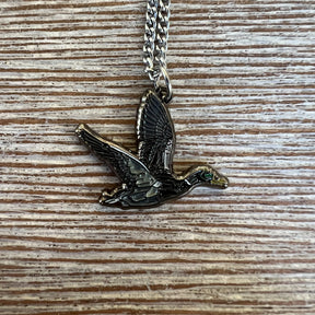 The Duck It OG Necklace by Mucker Bucket features a stainless steel flying bird pendant with detailed wings and body, set against a textured wood background and finished with a shiny surface. Proudly made in the USA.