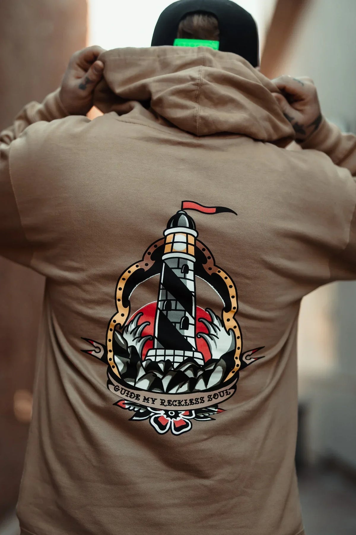 A person faces away in a camel Guide My Reckless Soul Hoodie by Relentless Betrayal, featuring a colorful lighthouse, waves, and red banner graphic with “Guide My Reckless Soul” text. Made from soft cotton-polyester fleece. Hood is up.