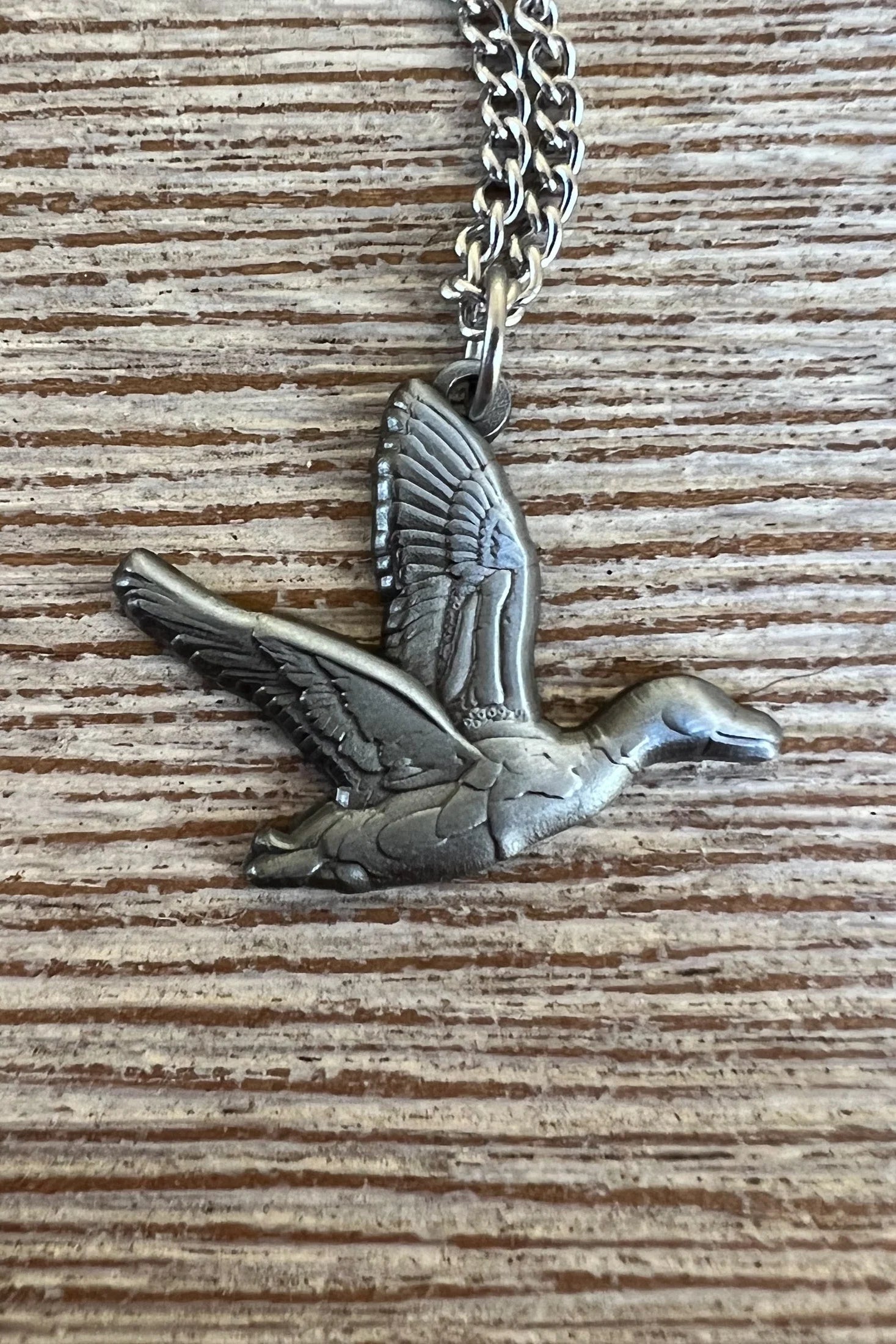 The Duck It Silver Necklace by Mucker Bucket features a flying bird pendant on a stainless steel chain, elegantly resting on a textured wooden surface. Its intricate design and graceful motion showcase quality craftsmanship. Made in the USA.