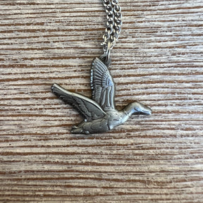 The Duck It Silver Necklace by Mucker Bucket features a flying bird pendant on a stainless steel chain, elegantly resting on a textured wooden surface. Its intricate design and graceful motion showcase quality craftsmanship. Made in the USA.