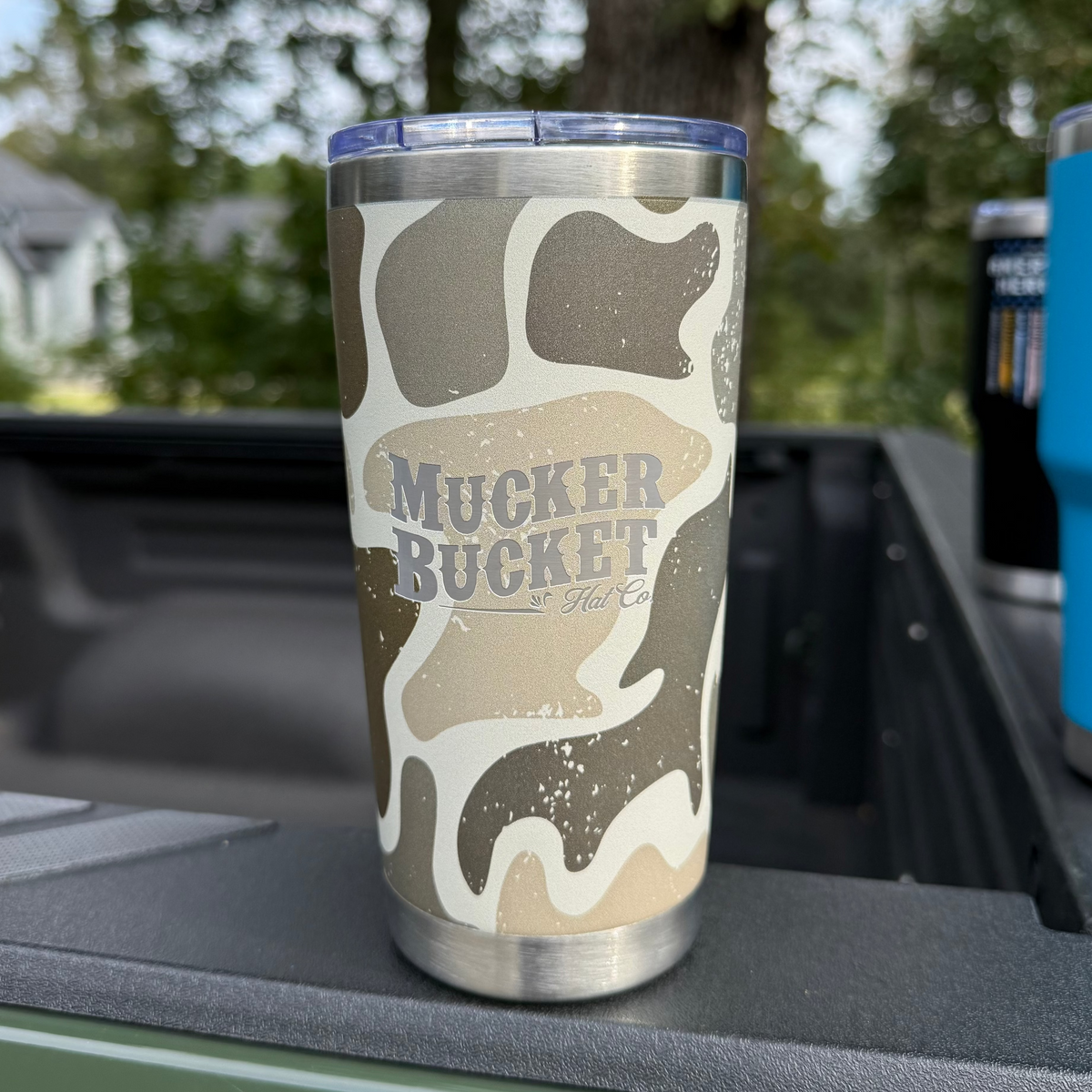 A 20oz Duck Camo Tumbler by Mucker Bucket, featuring a laser-engraved logo on stainless steel, rests on a truck bed with blurred trees and houses in the background.