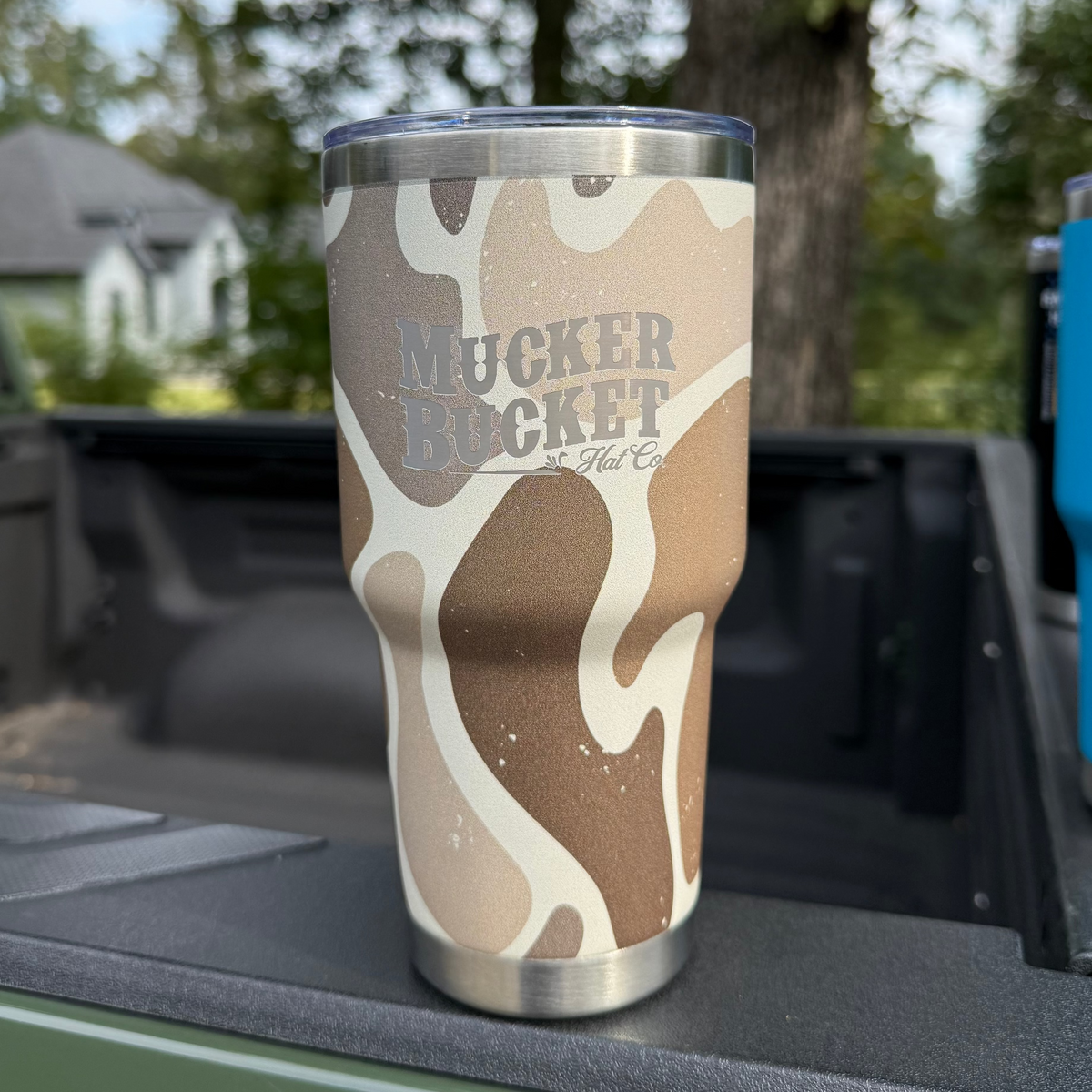 The Duck Camo Tumbler - 30oz by Mucker Bucket, featuring a tan and cream duck camo pattern and bold branding, rests on a truck bed outdoors with trees and a house in the background.