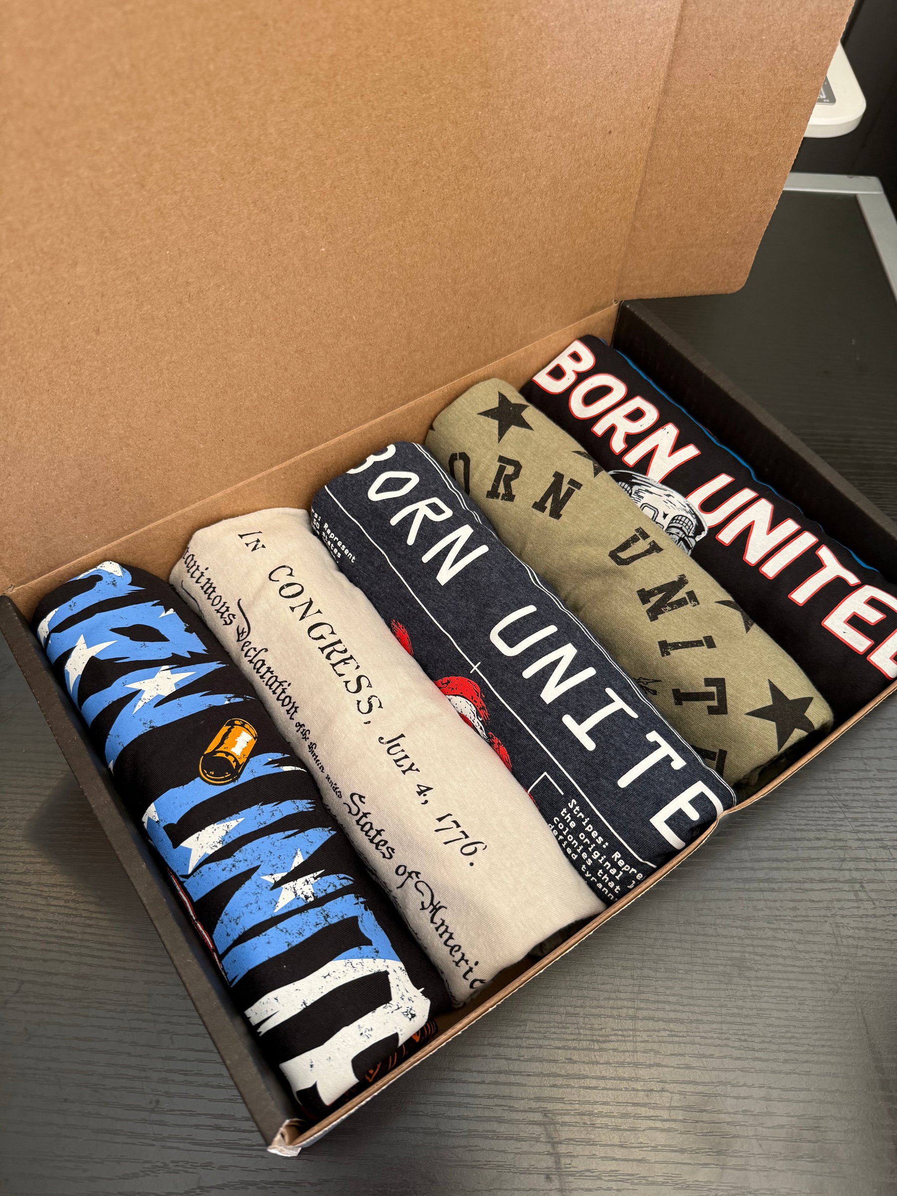 The Born United Patriot Box by Born United Private Label sits on a black surface and includes five folded t-shirts with patriotic designs—stars, stripes, and Constitution-inspired text. Features Limited Edition Tees ideal for collectors and enthusiasts.