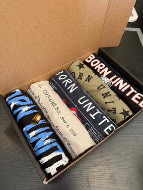 The Born United Patriot Box by Born United Private Label sits on a black surface and includes five folded t-shirts with patriotic designs—stars, stripes, and Constitution-inspired text. Features Limited Edition Tees ideal for collectors and enthusiasts.