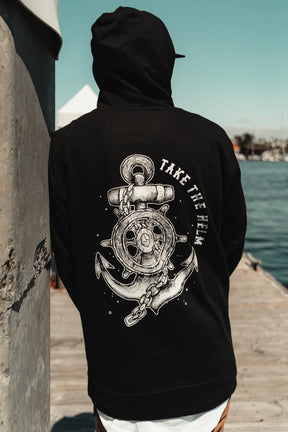 A person wearing the Relentless Betrayal Take The Helm Black Hoodie, featuring a nautical anchor and ship wheel graphic with "TAKE THE HELM," stands on a dock by the water, facing away from the camera.