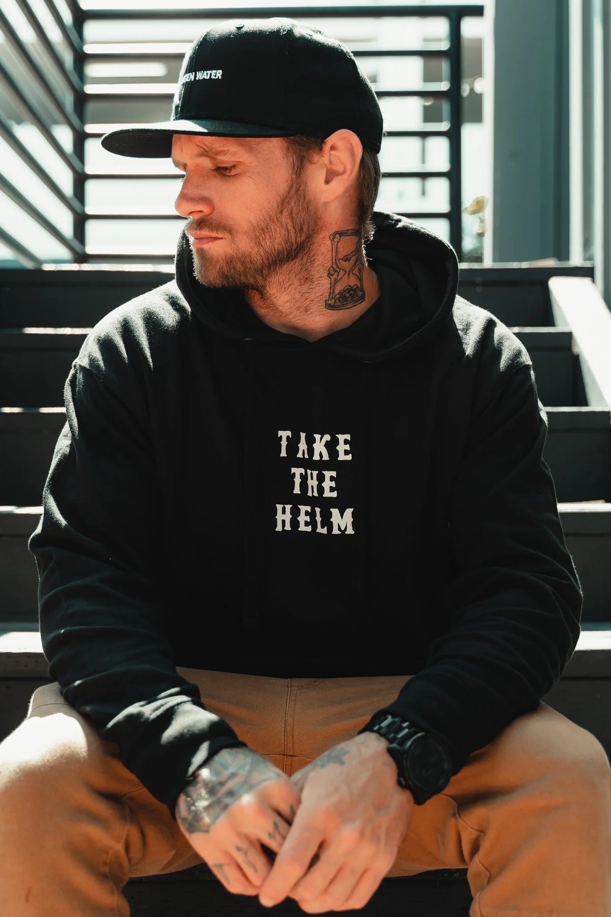 A tattooed man sits on outdoor steps, wearing the Relentless Betrayal Take The Helm Black Hoodie and a black cap. He looks to the side with hands on his knees, capturing the spirit of this inspirational apparel in soft sunlight.