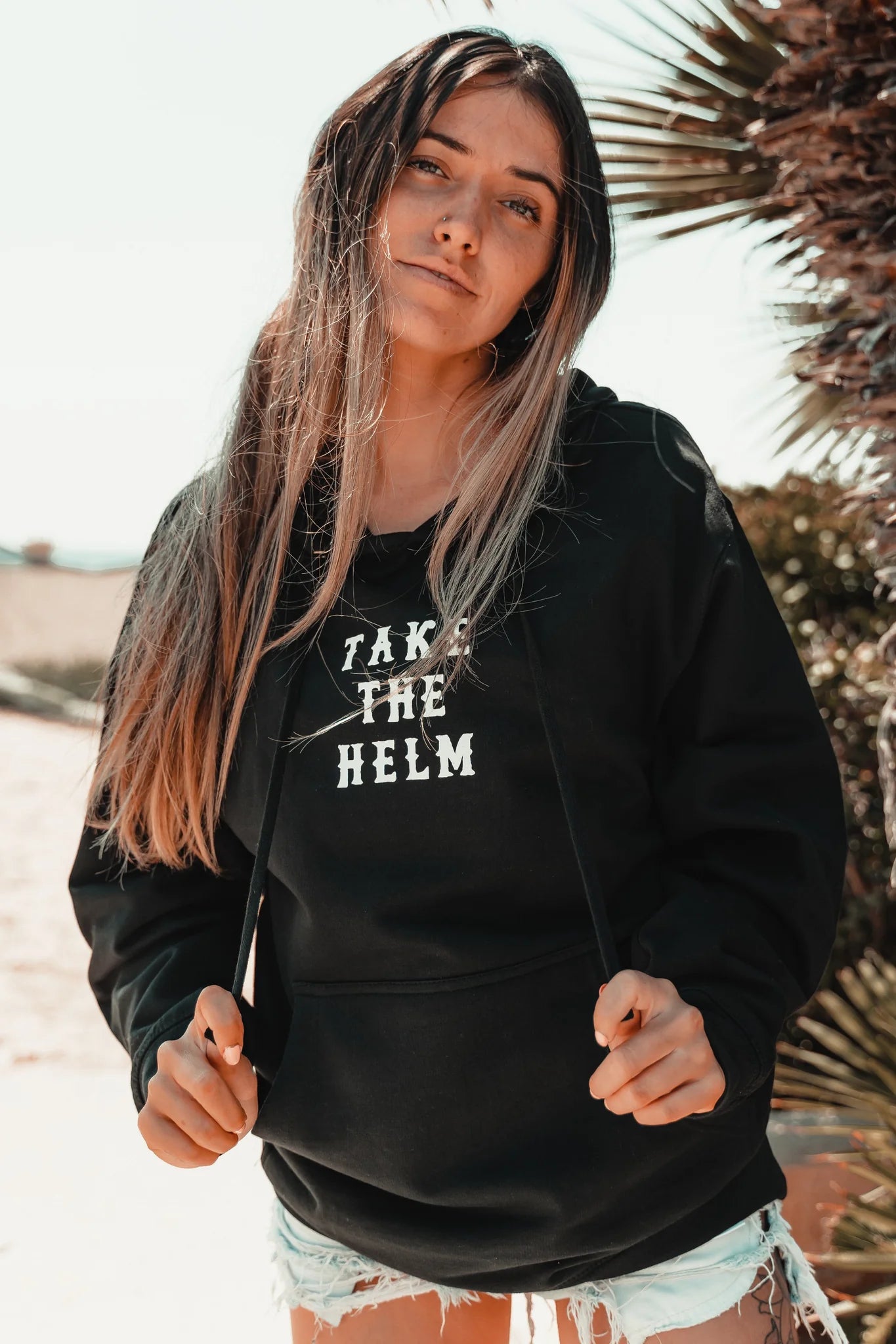 A young woman with long hair wears the Relentless Betrayal Take The Helm Black Hoodie and distressed denim shorts, standing outdoors by palm trees in bright sunlight.