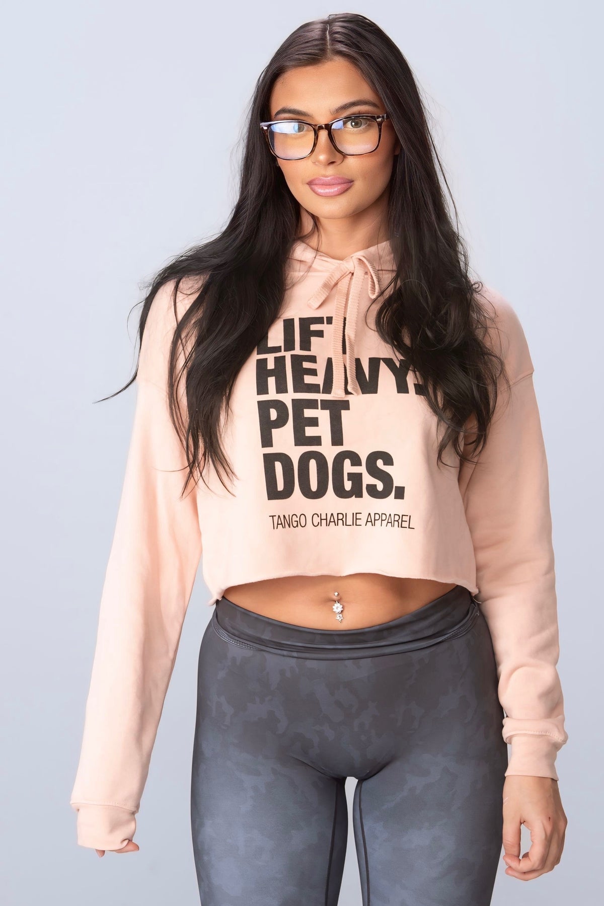 A woman with long dark hair and glasses wears a comfy "Lift Heavy. Pet Dogs." crop hoodie by Tango Charlie Apparel, paired with gray high-waisted leggings. She has a belly button piercing and stands against a light background.