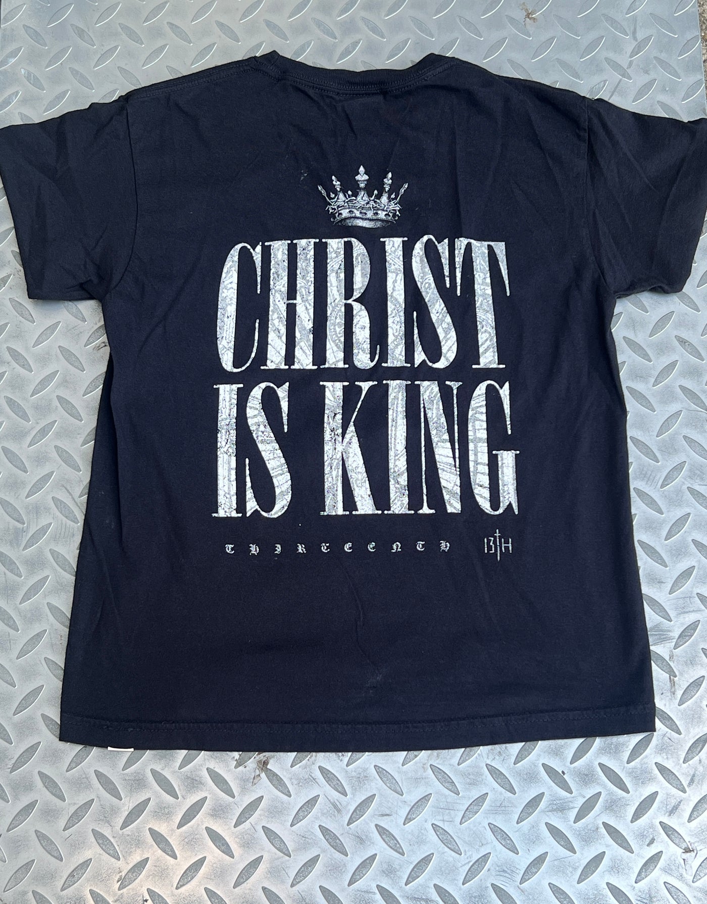 The Youth Christ is King Tee by 13th features a crown above bold white CHRIST IS KING text and smaller THIRTEEN branding with the 13th logo, shown laid flat on a metal-patterned surface.