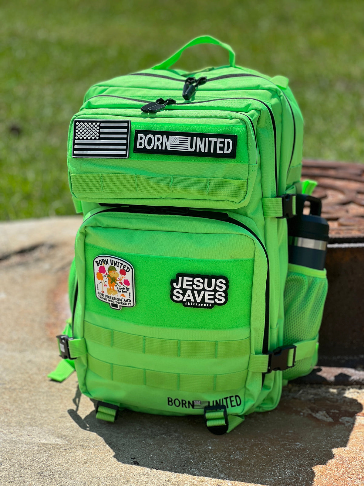 The Key Lime Tac-Pac - 45L by Born United Private Label, shown outdoors with “BORN UNITED,” “JESUS SAVES,” and flag patches, plus a round graphic patch and a water bottle in the side pocket, sits on concrete.