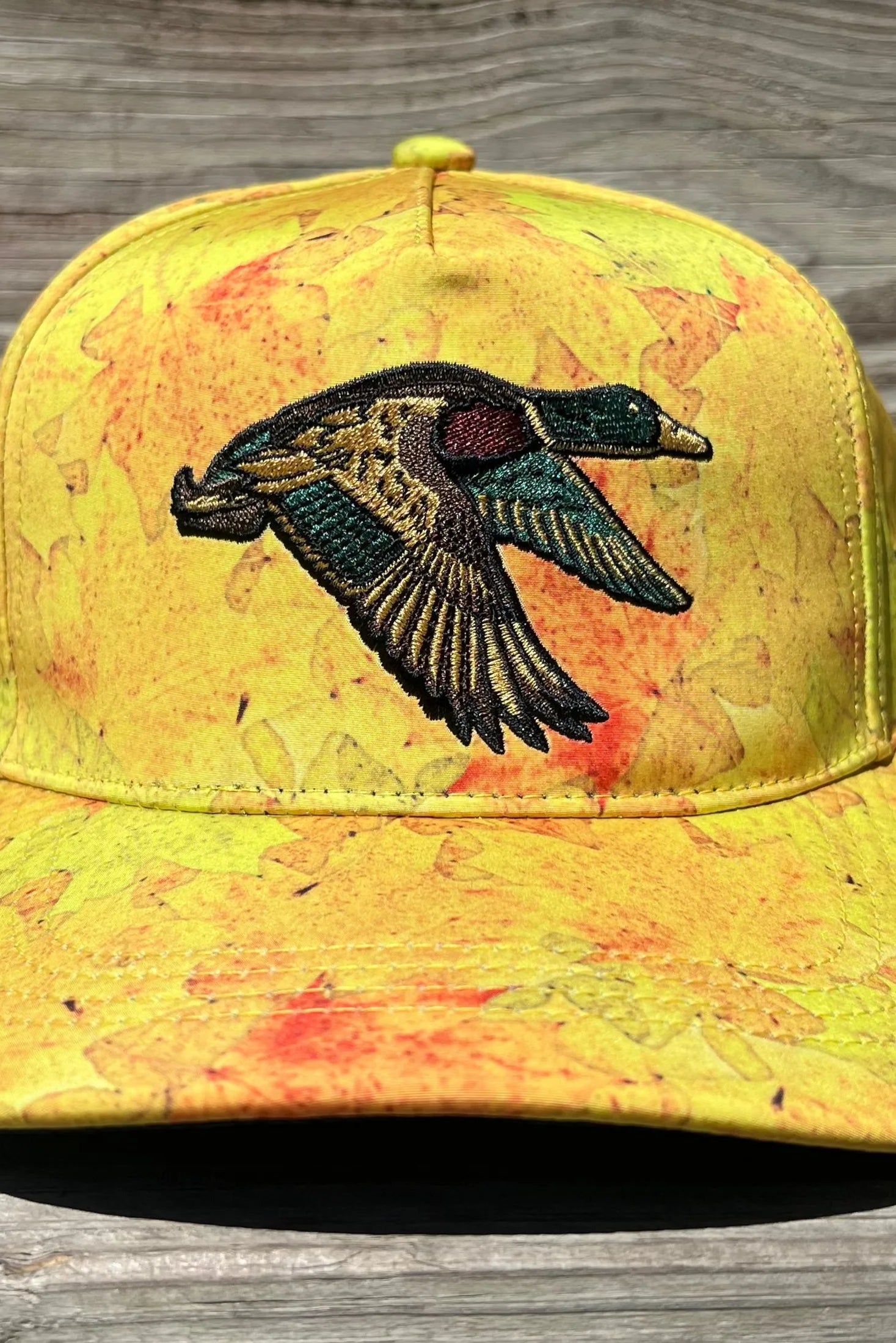 The Golden Leaf Camo Duck Bucket by Mucker Bucket exudes duck hunting charm, featuring a yellow camo leaf pattern and an embroidered duck in flight on the front. Its displayed on a wooden surface to highlight the standout design.