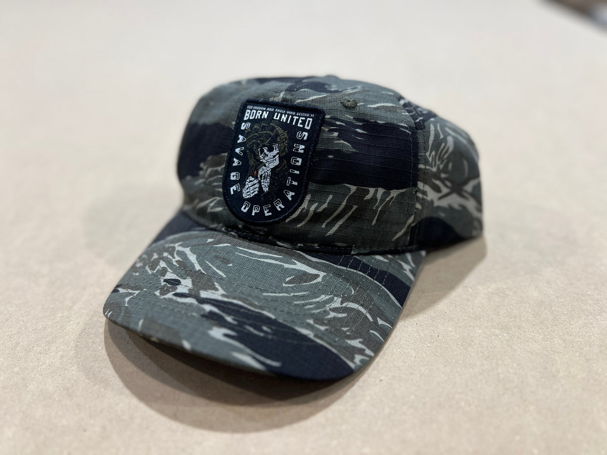 The Born United Private Label "Born United Buckle Hat - Savage Operations," a camo baseball cap featuring an eagle emblem and “BORN UNITED WAR OPERATIONS” patch, is displayed on a light-colored surface.