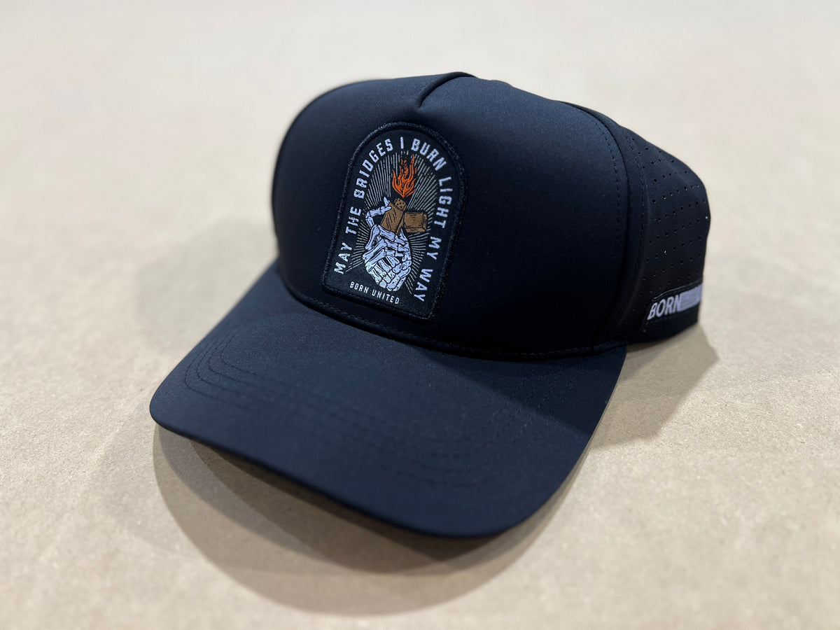 The Born United Premium Snapback - Burning Bridges by Born United Private Label features a black design with a burning torch patch and "May the bridges I burn light my way" embroidered on the front, plus a side label. Displayed on a beige surface.