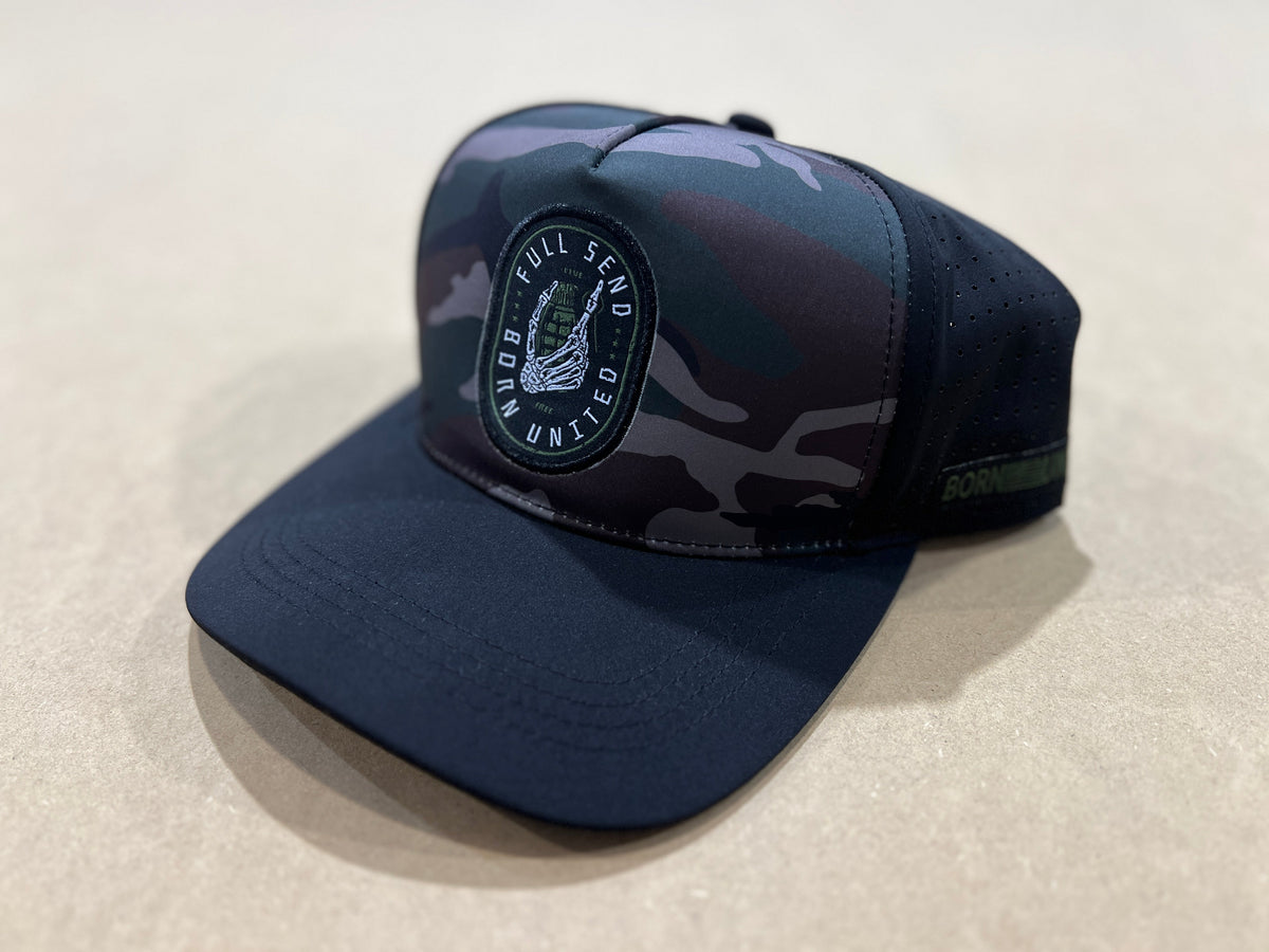 The Born United Premium Snapback - Full Send (Camo) by Born United Private Label is a black and gray camo trucker hat featuring a round patch with an eagle emblem on the front, shown on a beige surface.