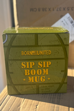 A green box labeled with the Born United Private Label brand and “BU Sip Sip Boom Mug” in yellow letters rests on cardboard, with a shipping label partly visible in the background.