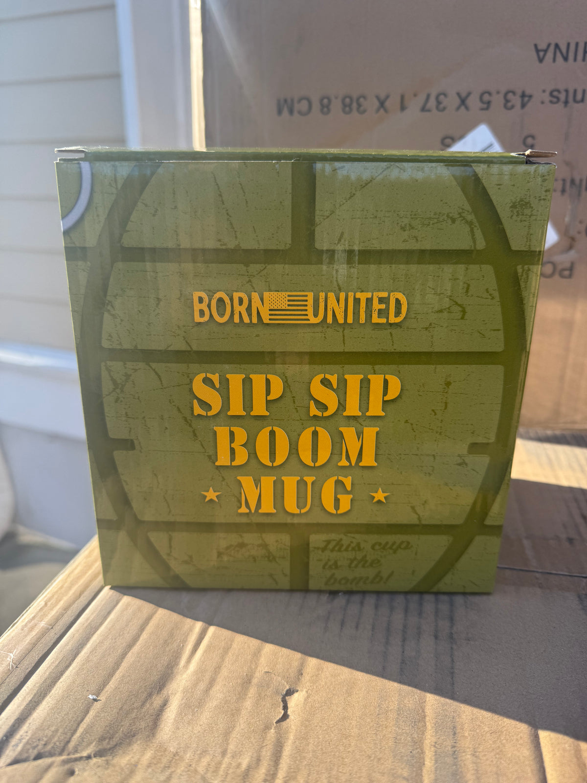 A green Born United Private Label BU Sip Sip Boom Mug sits outdoors in sunlight, featuring a subtle grenade pattern and cardboard boxes in the background.