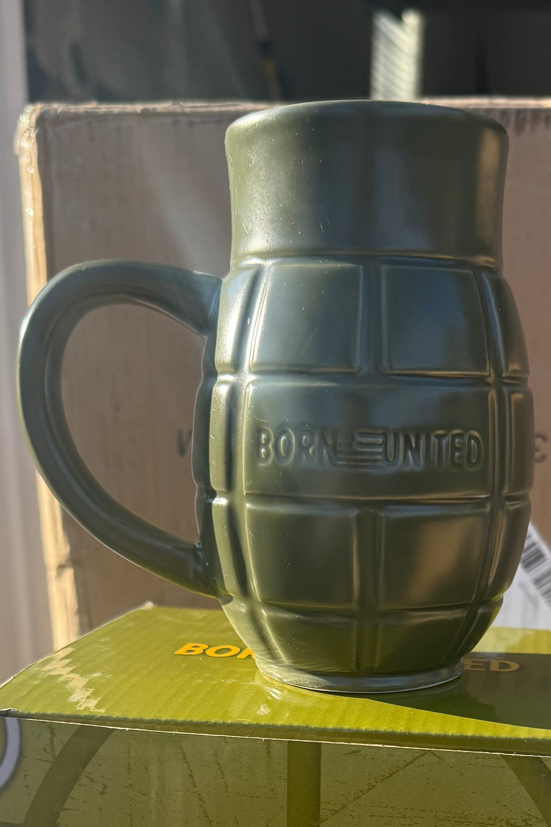 The BU Sip Sip Boom Mug by Born United Private Label is a green ceramic hand grenade-shaped mug with "BORN UNITED" embossed on its side, displayed on a box with sunlight casting shadows.