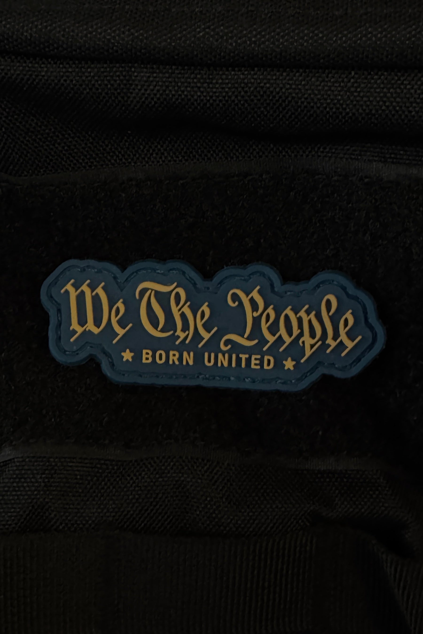 A bold 3-inch “We The People PVC Patch” by Born United Private Label features gold script reading “We The People BORN UNITED” on a dark, star-studded background, and attaches securely to black fabric with Velcro.
