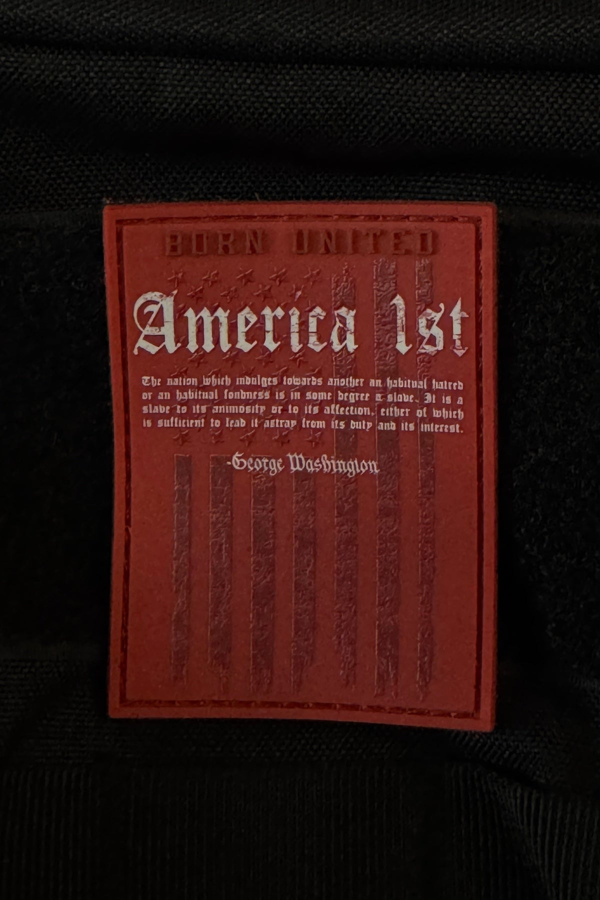 A striking black PVC patch by Born United Private Label, measuring 3 inches and equipped with Velcro, displays a bold red label featuring the text “America First,” a George Washington quote, a faded American flag motif, and “Born United” at the top.