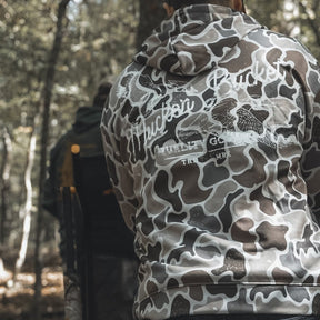 A person wearing a gray and white Mucker Bucket Wood Ducks Premium Athletic Duck Camo Hoodie with "Huckin Bucket" on the back sits outdoors in a wooded area, while another person is seated in the background.