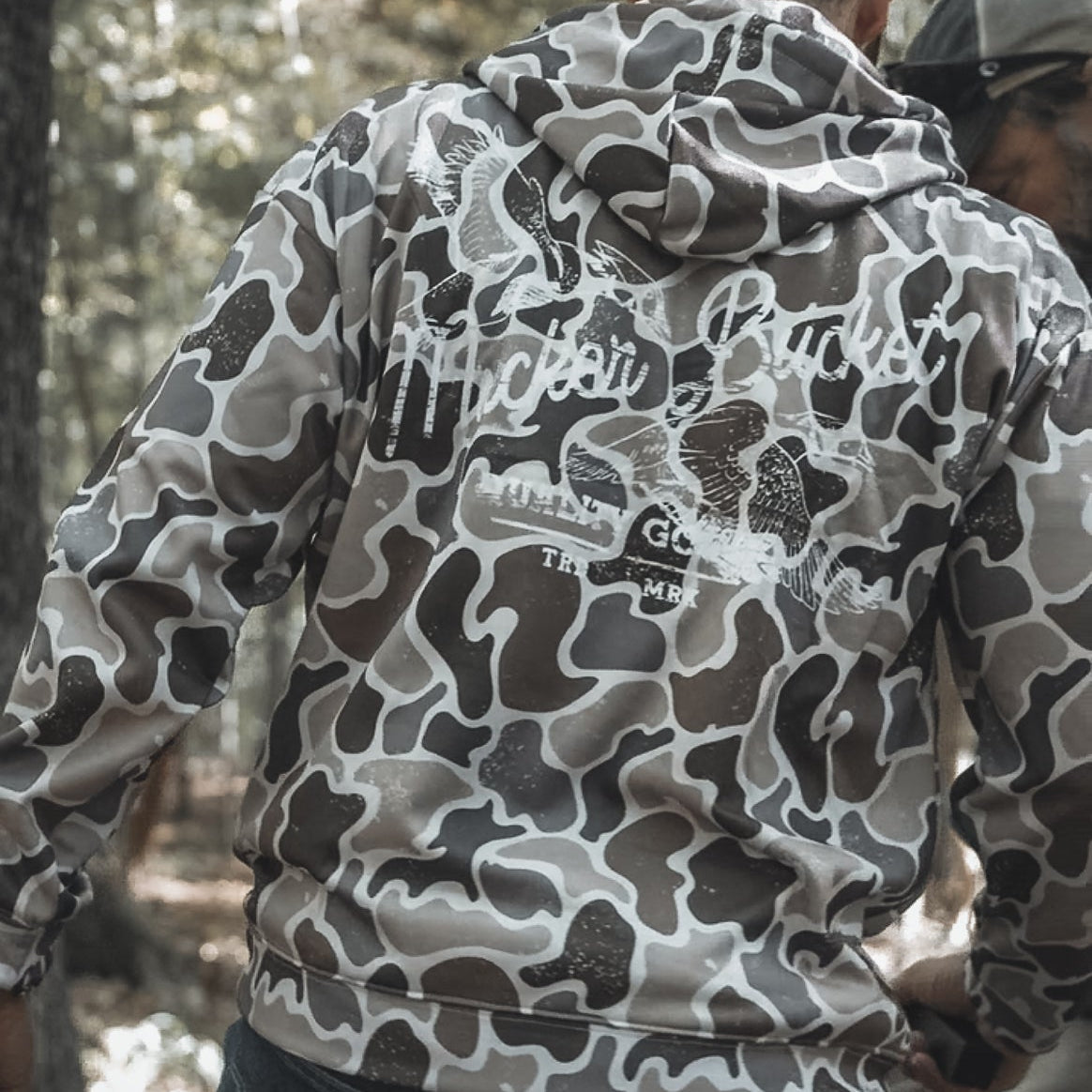 Someone wearing the Mucker Bucket Wood Ducks Premium Athletic Duck Camo Hoodie, featuring “Hacker Cracker” on the back, stands outdoors in a wooded area with their back to the camera.